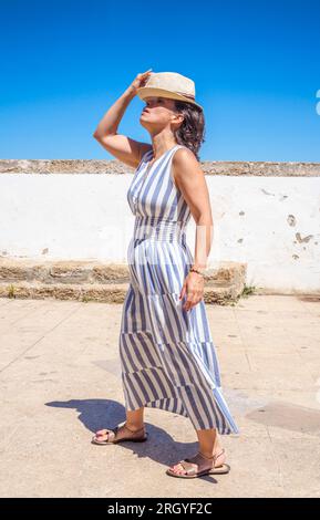 Smiling Caucasian tourist wearing a summer dress and a vintage straw hat, walking an antique Latinamerican marina promenade. Stock Photo