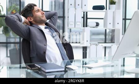 Taking time for a minute break. Cheerful young man holding hands behind head and keeping eyes closed while sitting at his working place Stock Photo