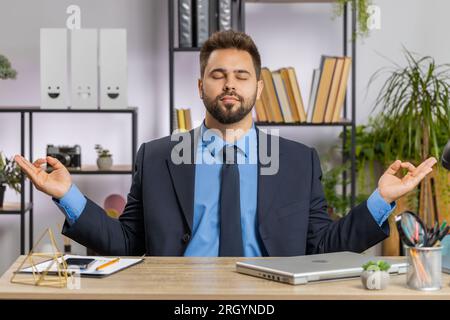 Caucasian businessman working on laptop computer, meditating, doing yoga breathing exercise in lotus position at home office. Calm serene freelancer man taking break. Busy occupation. Peace of mind Stock Photo