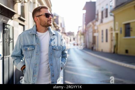 A handsome young adult brunette in sunglasses and a denim jacket walks around the city. Stock Photo