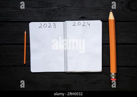 The year 2023 and 2024 is written on the pages of a notebook and next to it lies a large and small pencil on a black table, the end of the old year an Stock Photo