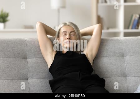 Calm blonde mature lady relaxing on grey comfortable home sofa Stock Photo
