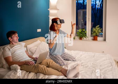 Woman enjoys virtual reality with VR headset, smiling admiringly in company of her husband Stock Photo