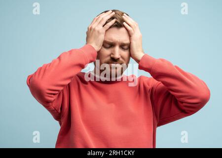 Portrait of sad man having life troubles holding head with suffering face on studio blue background. Stock Photo
