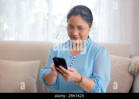 Asian elderly woman using smartphone to social media online sitting on sofa with relax in living room at home, senior woman looking smart phone on cou Stock Photo