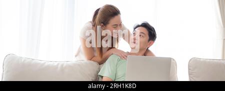 Young asian couple using laptop computer for looking social media or video with relax and leisure on sofa in the living room at home, man and woman wa Stock Photo