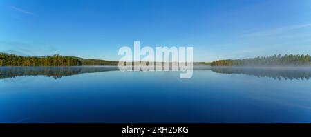 The mirror-like tranquil lake surface reflects the reflection of woods and mountains. Stock Photo