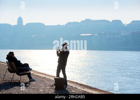 Silhouette of man taking photo of woman sitting on the bench at the river against city skyline Stock Photo