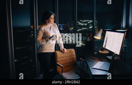businesswoman working late in her office, engaged in planning a social media marketing project. She stands thoughtfully in front of her desk, holding Stock Photo