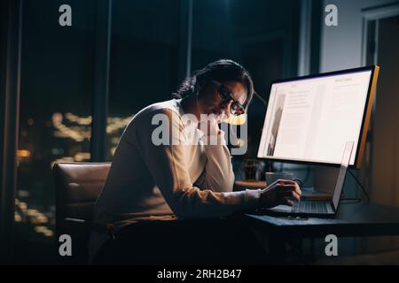 business woman experiencing workplace burnout and computer fatigue while working late at her dimly lit office desk. Woman feeling exhausted as she wor Stock Photo