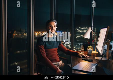 A caucasian male entrepreneur works late at his office desk, conducting market research with his laptop. He sits confidently, looking at the camera, s Stock Photo