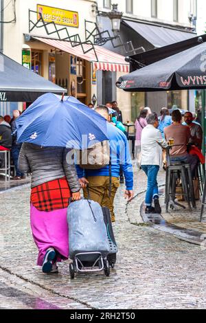 Couple with roller baggage walking along city street under on eumbrella during rainstorm - Brussels, Belgium. Stock Photo