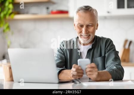 Smiling Senior Man Using Smartphone And Laptop Computer At Home Stock Photo