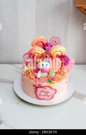 Birthday cake with cute fondant unicorn, lollipos, meringues, popsicles and chocolate flowers for a girl or a baby. Text "Happy Birthday" Stock Photo