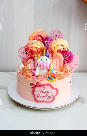 Birthday cake with cute fondant unicorn, lollipos, meringues, popsicles and chocolate flowers for a girl or a baby. Text "Happy Birthday" Stock Photo