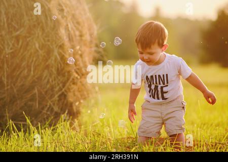 The child, a boy, is playing on the field with freshly cut grass, surrounded by hay stacks, against the backdrop of a sunset. Portrait of a happy kid Stock Photo