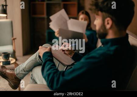 Two businesspeople working, the man is writing something on his note book and the woman is reading through the paper documents with the laptop in her Stock Photo