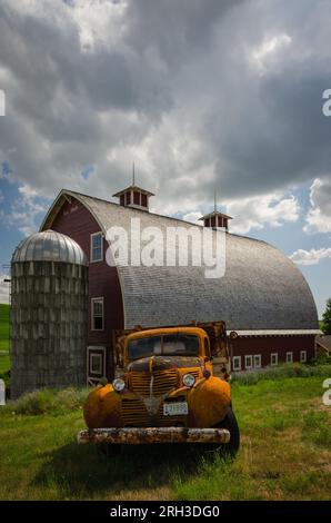 A rusty old orange truck and a large red barn (built 1919) with cedar roof, two cupolas and a small silo. Colfax, Washington, USA. Stock Photo