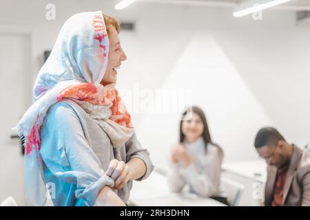 Redhead muslim businesswoman giving presentation speech to a multiracial group of businesspeople. Ginger businesswoman wearing hijab listening to a co Stock Photo