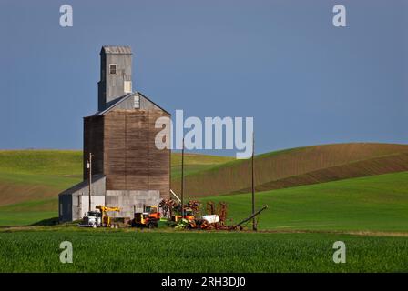 Old grain elevator and a variety of farm equipment, surrounded by rolling wheat fields in spring. Whitman County, Washington, USA. Stock Photo