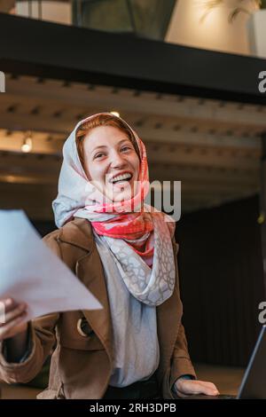Cheerful muslim female entrepreneur in hijab holding document and smiling Stock Photo