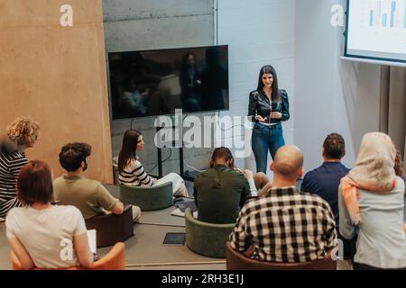 Brunette female speaker giving speech to multiracial group of entrepreneurs, using presentation, tv screen, graph charts, statistics. Business people Stock Photo