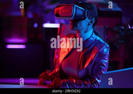 Neon metaverse futuristic concept. modern 40 years old woman in virtual reality goggles in modern office. Stock Photo