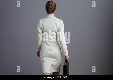 Seen from behind female employee in a light business suit with briefcase walking against gray background. Stock Photo