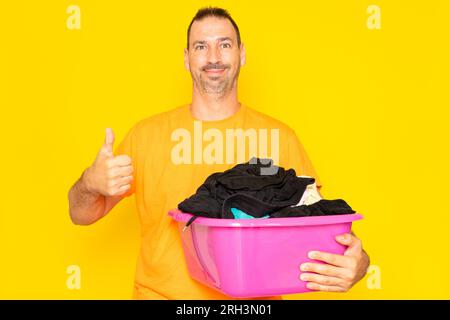 Bearded hispanic man doing housework holding plastic basket with clothes raising thumb up, excellent symbol. Isolated on yellow background. Stock Photo