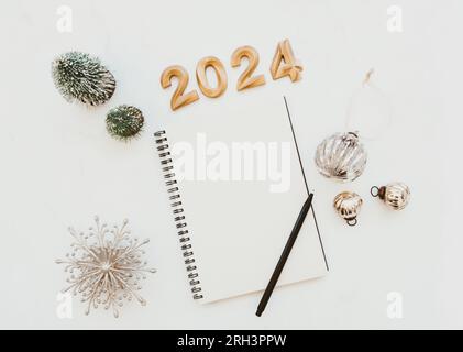 Flat lay of blank notebook sheet with copy space, numbers 2024, christmas decoration, christmas balls on white marble background. Concept planning, wi Stock Photo