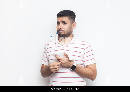 Portrait of sick sad unhealthy bearded man wearing striped t-shirt standing with thermometer in his mouth, having grippe symptoms, looking at camera. Indoor studio shot isolated on gray background. Stock Photo
