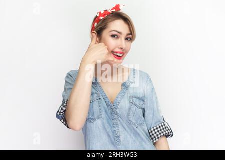 Call me back. Portrait of flirting joyful attractive blonde woman wearing blue denim shirt and red headband standing showing call me gesture. studio shot isolated on gray background. Stock Photo