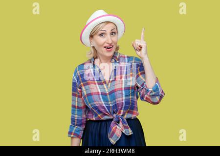 Portrait of senior woman wearing checkered shirt and hat get new idea, raises fore finger, ready to start work on project, confident in success. Indoor studio shot isolated on yellow background. Stock Photo