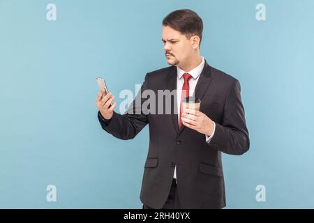 Portrait of serious concentrated man with mustache standing using mobile phone and drinking takeaway coffee, wearing black suit with red tie. Indoor studio shot isolated on light blue background. Stock Photo