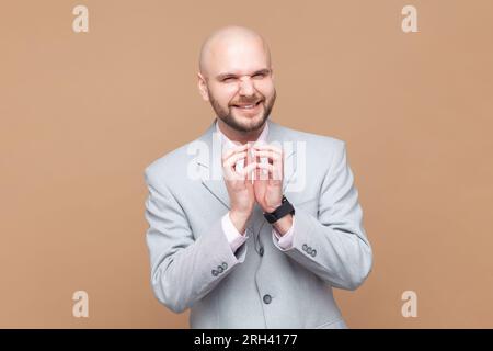Portrait of sly smiling bald bearded man looking at camera with cunning facial expression, planning devil prank, wearing gray jacket. Indoor studio shot isolated on brown background. Stock Photo