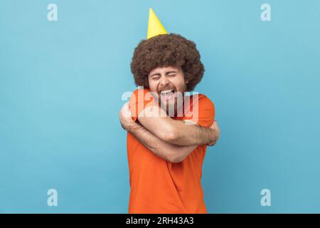 Man with Afro hairstyle in party cone embracing himself with expression of pleasure, happy to celebrate holiday alone, self-love concept. Indoor studio shot isolated on blue background. Stock Photo