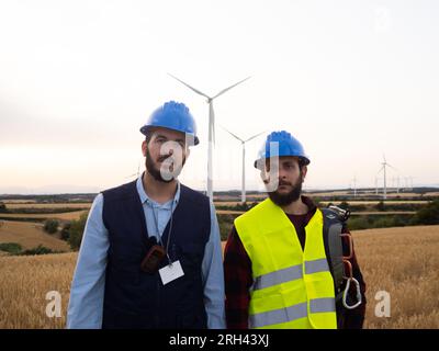Two male industrial engineer workers looking seriously at camera in a field of wind mills Stock Photo