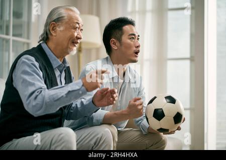 asian senior father and adult son watching football match on tv together Stock Photo