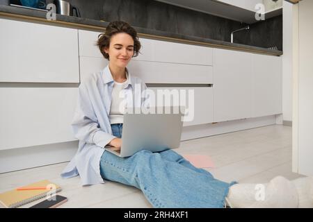 Portrait of girl freelancer, sits at home on floor and works with laptop, surrounded with piles of documents, smiles while looks at computer screen Stock Photo