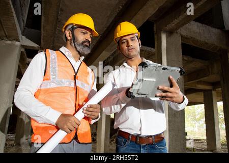Two Indian male civil engineers or architect wearing helmet and vest holding digital tablet blueprint at construction site discussing real estate proj Stock Photo