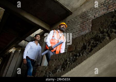 Two Indian male civil engineers or architect wearing helmet and vest holding paperwork blueprint walking on stairs of under construction building and Stock Photo