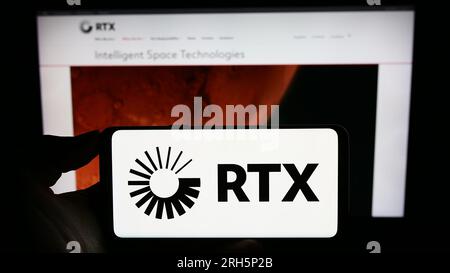 Person holding cellphone with logo of US aerospace and defense company RTX Corporation on screen in front of webpage. Focus on phone display. Stock Photo