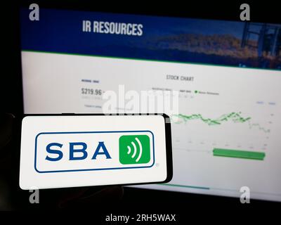 Person holding mobile phone with logo of American company SBA Communications Corporation on screen in front of web page. Focus on phone display. Stock Photo