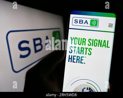 Person holding cellphone with webpage of US company SBA Communications Corporation on screen in front of logo. Focus on center of phone display. Stock Photo