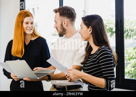 Diverse, ecstatic businesspeople discussing papers and working on laptops at an office table while smiling Stock Photo
