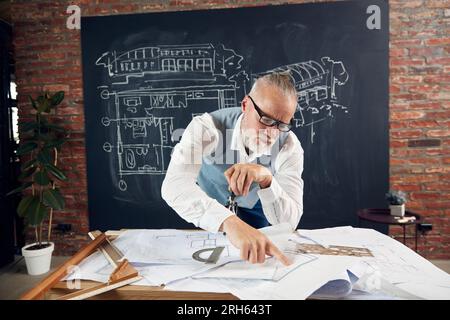 Bearded senior man, architect sitting at table and working with buildings design, sketches. Making measurements Stock Photo