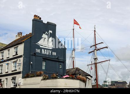 The side of the Navy Inn in Southside Street on Plymouth’s Barbican. Its logo above its rooftop beer garden reflected by the mast of the Oosterschelde Stock Photo