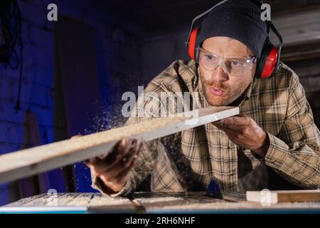 carpenter blowing sawdust from wooden plank after cutting with circular saw. man working in workshop Stock Photo