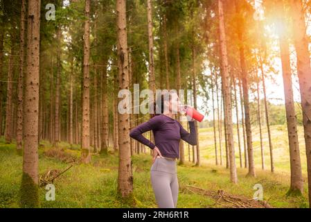 Sporty woman taking a drink from a glass bottle, exercise in the forest. Stock Photo