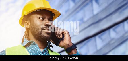 banner of male civil engineer of African ethnicity wearing yellow safety helmet and reflective vest, outdoors next to building, talking on phone, copy Stock Photo